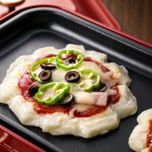 Mochi pizza with ketchup, cheese, green pepper, bacon and black olives on a hot plate